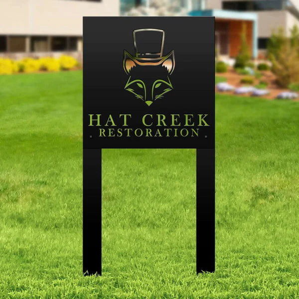 Black metal sign with fox in top hat and “HAT CREEK RESTORATION,” mounted on posts in grassy landscaped area.