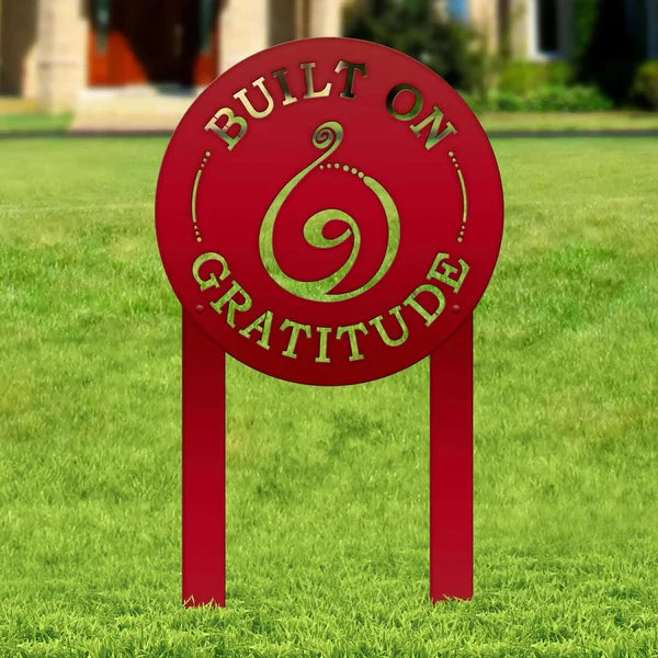 Deep red black laser-cut sign with “BUILT ON GRATITUDE” and spiral cutout, mounted on posts in grass lawn with house behind.