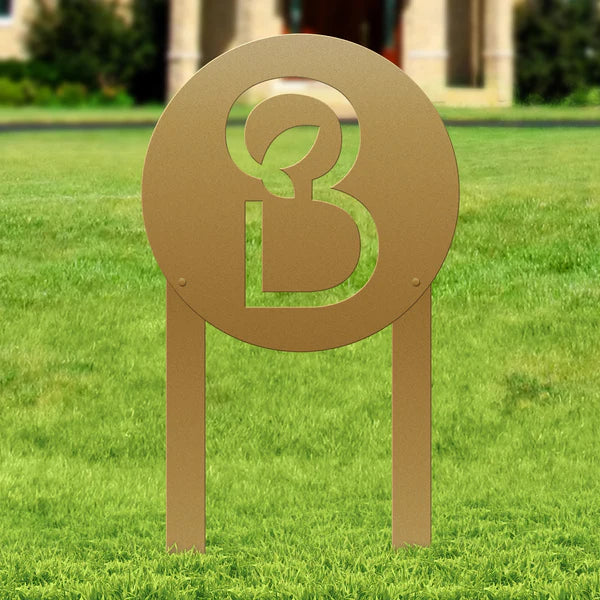 Laser-cut gold metal sign with “B” and leaf in circular frame, mounted on two legs, placed in grassy outdoor area.