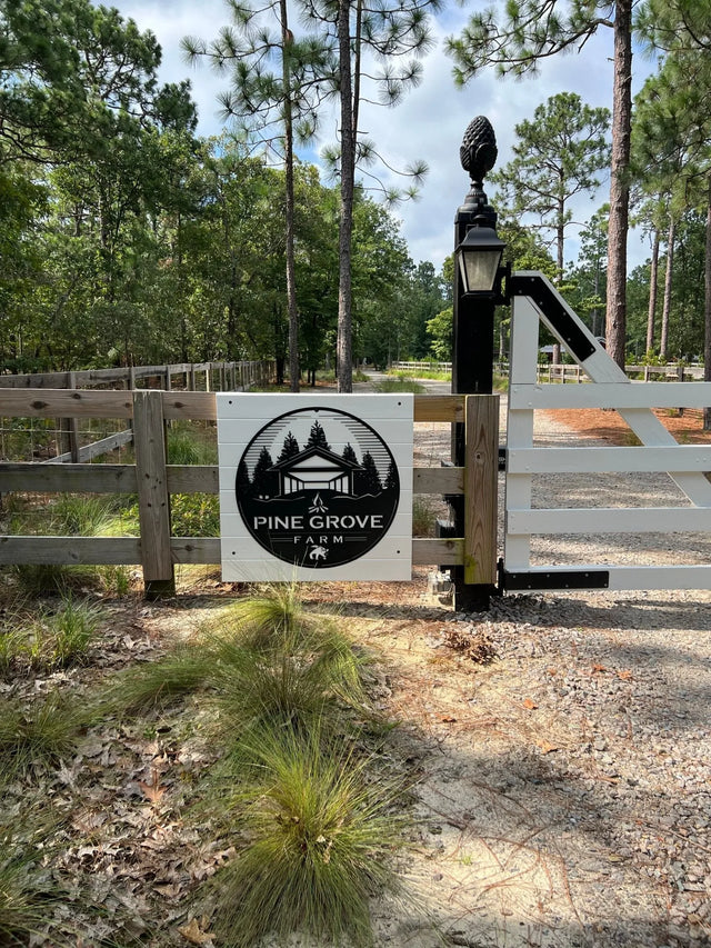 Metal sign on wood fence says “Pine Grove Farm,” with house and tree illustration set against pine forest background.
