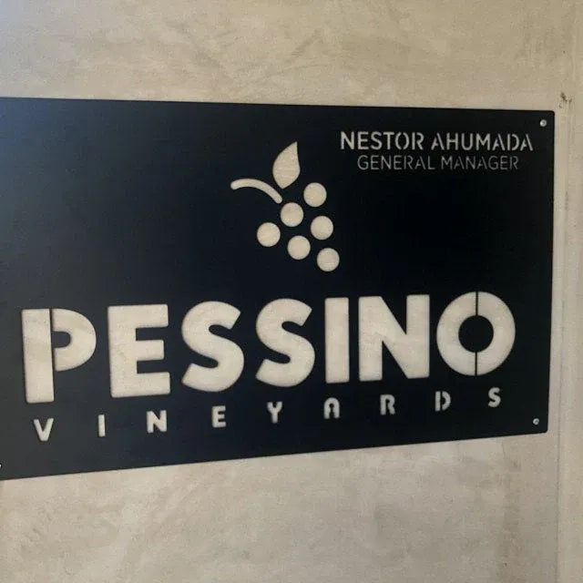 Laser-cut sign with bold “PESSINO,” vertical “VINEYARDS,” grape cluster, and “NESTOR AHUMADA GENERAL MANAGER” at top left.