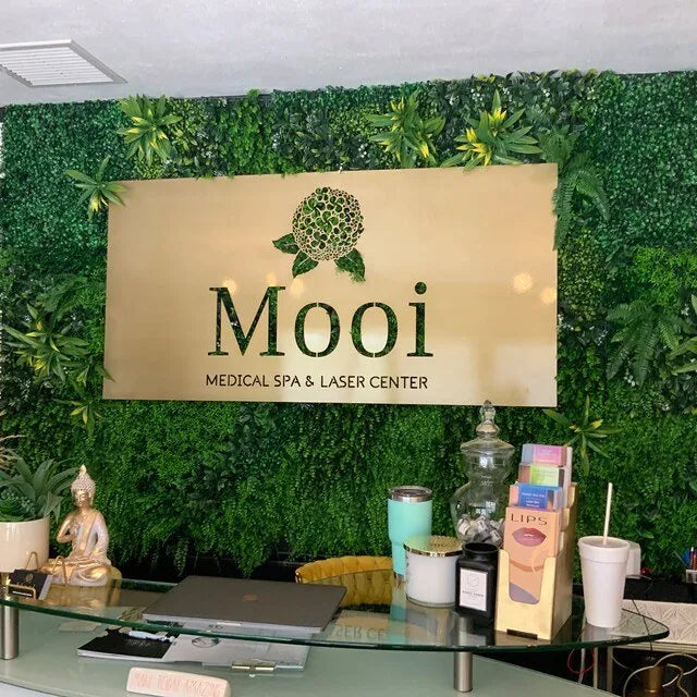 Reception area of “Mooi” Medical Spa & Laser Center with floral logo sign on greenery wall above glass counter.