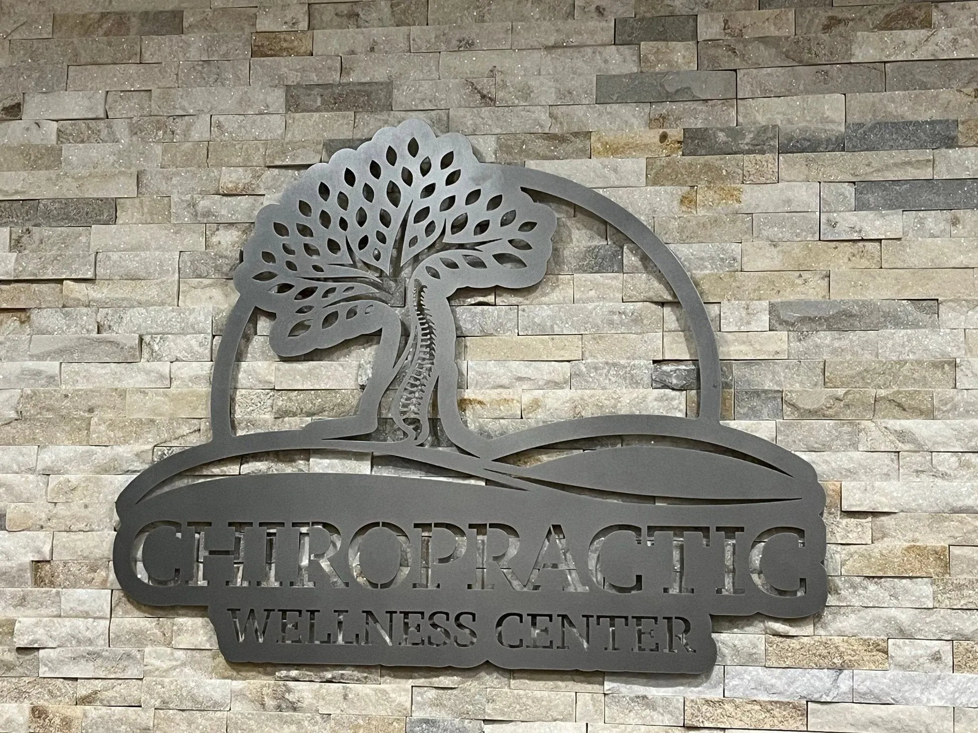 Laser-cut metal sign on stone-tile wall with vertical “CHIROPRACTIC WELLNESS CENTER” and circular tree-spine emblem.