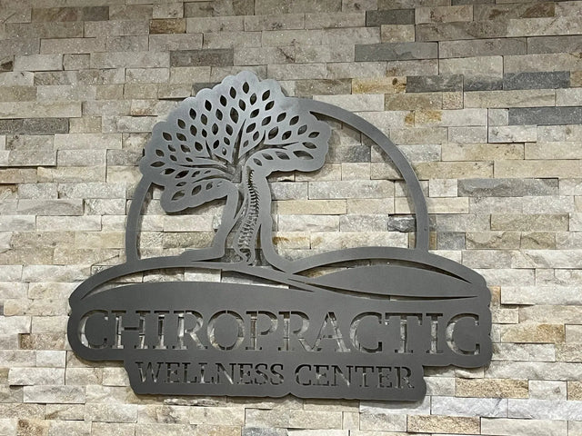 Laser-cut metal sign on stone-tile wall with vertical “CHIROPRACTIC WELLNESS CENTER” and circular tree-spine emblem.