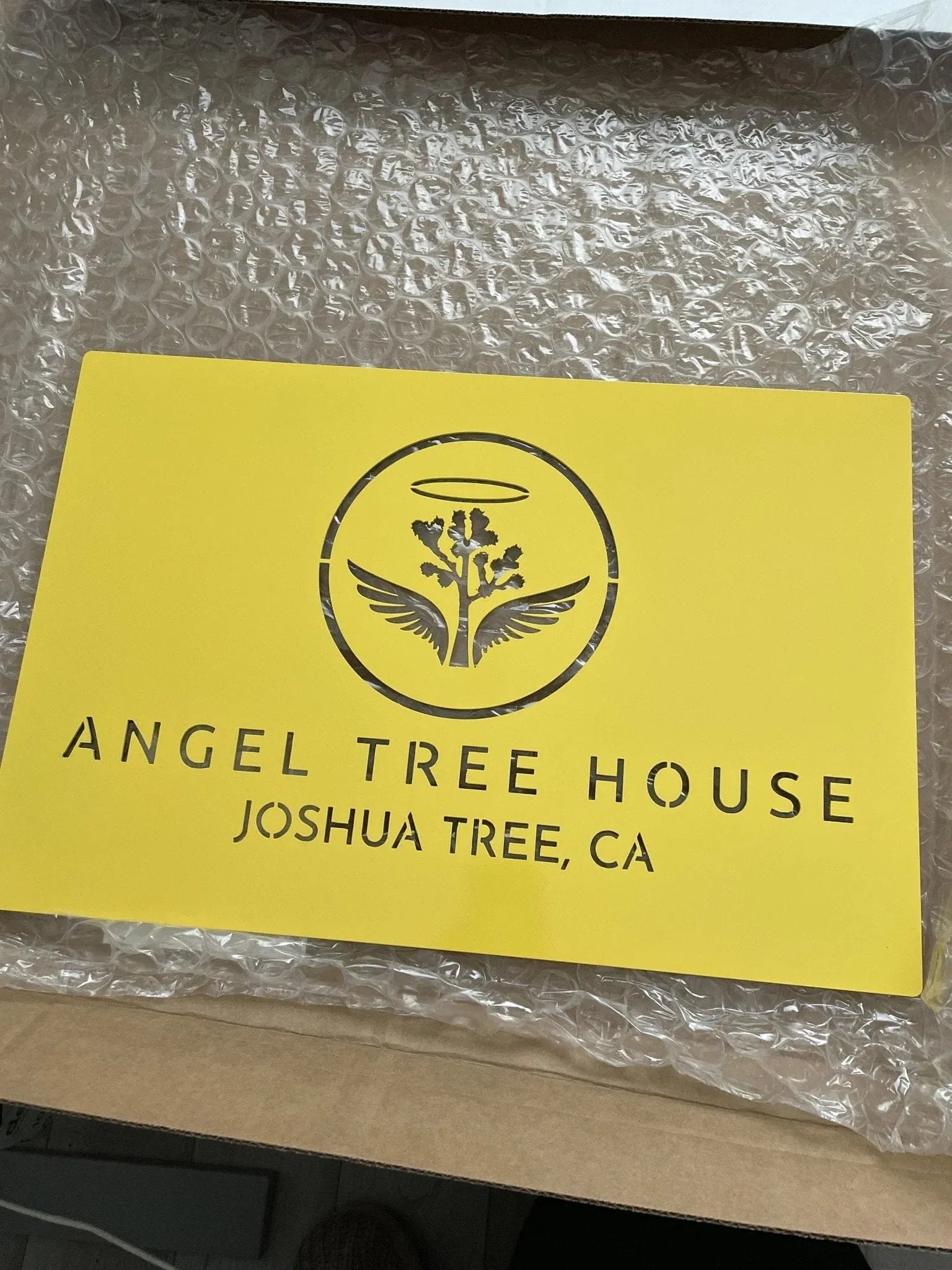 Yellow laser-cut steel sign with vertical “ANGEL TREE HOUSE” and “JOSHUA TREE, CA,” tree with wings and halo on bubble wrap.