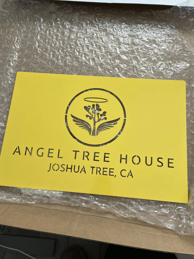 Yellow laser-cut steel sign with vertical “ANGEL TREE HOUSE” and “JOSHUA TREE, CA,” tree with wings and halo on bubble wrap.