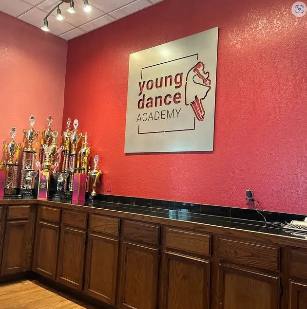 Laser-cut sign reads “young dance ACADEMY” with dancer silhouette, mounted on red wall above trophy counter.