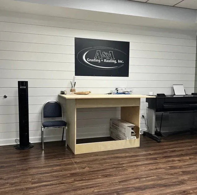 Wall-mounted black sign says “A&A Grading & Hauling, Inc.” in bold letters above wood desk on horizontal paneled wall.