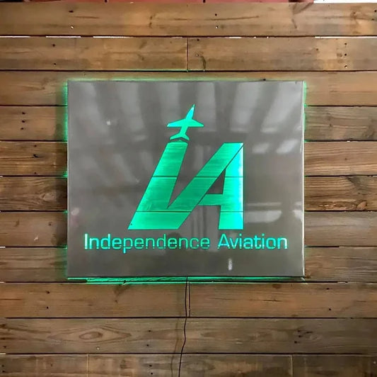 Green backlit 3D sign on wood wall: “Independence Aviation” with “IA” airplane logo and glowing edges.