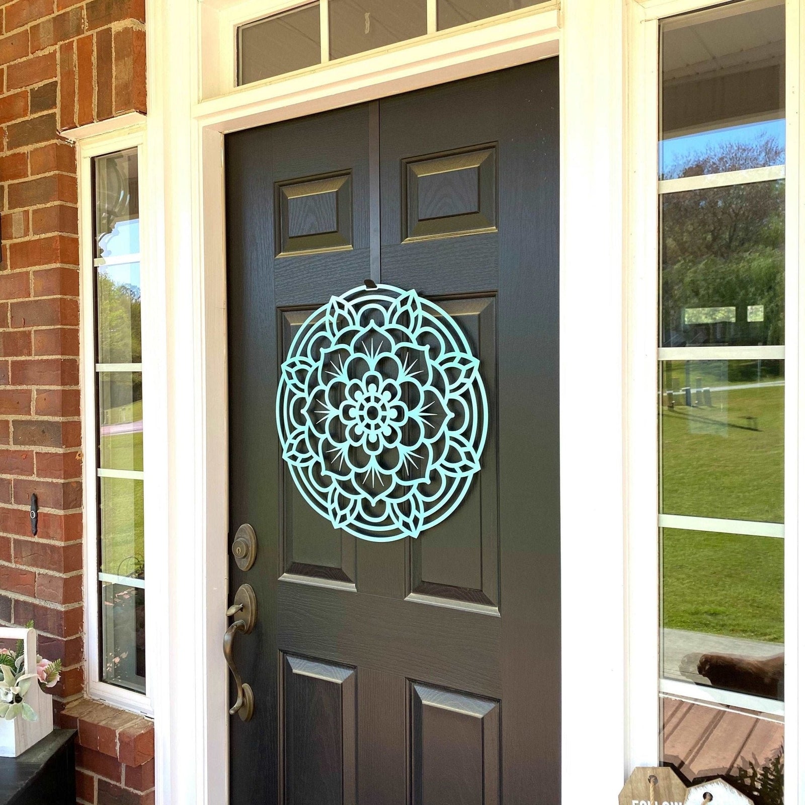 Decorative mandala wall art displayed on a gray front door, with a brick wall and glass windows in the background, featuring intricate lotus-inspired design and durable metal craftsmanship.