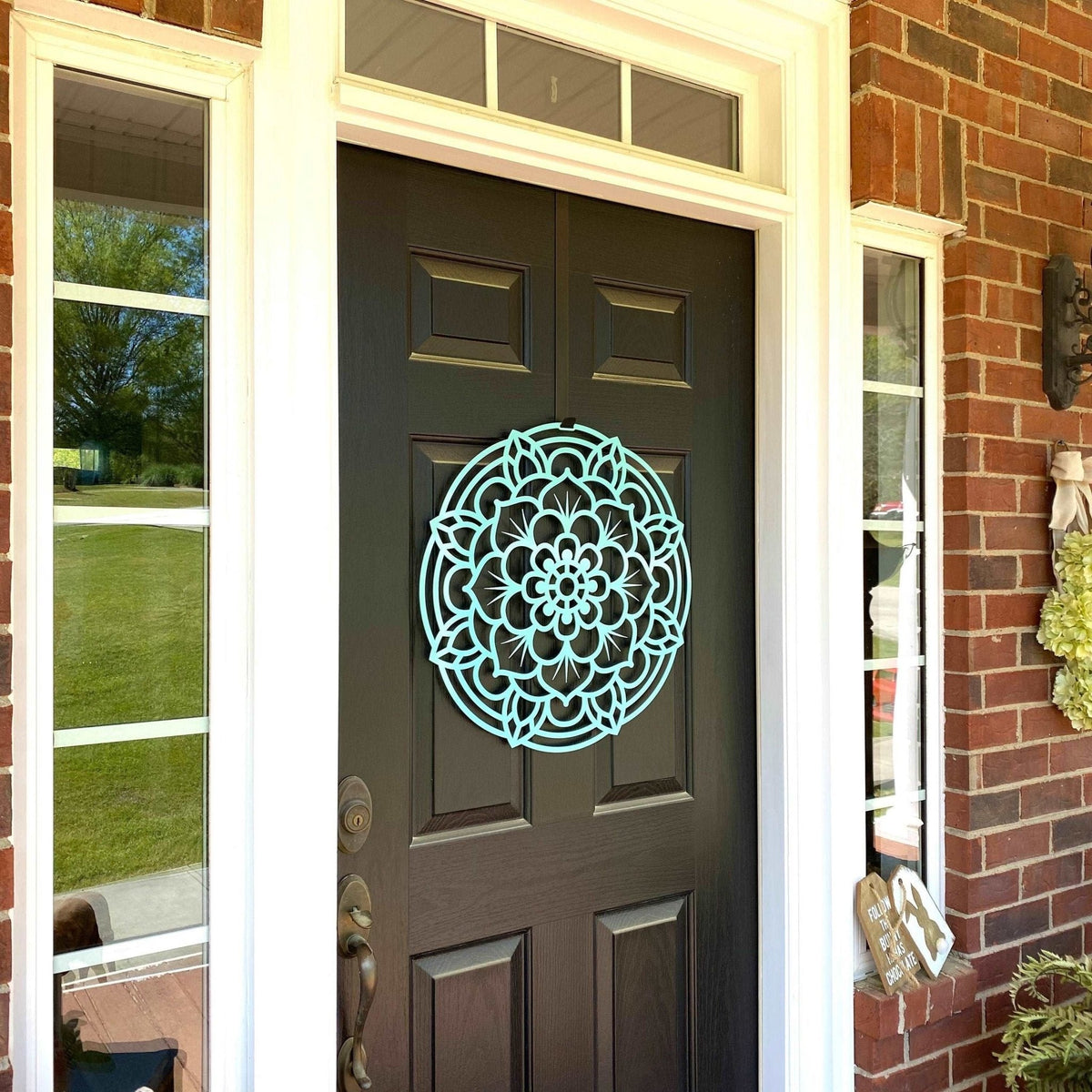 Decorative mandala wall art displayed on a dark brown front door, mounted on a brick wall with windows in the background, featuring an intricate lotus-inspired design and durable metal craftsmanship.