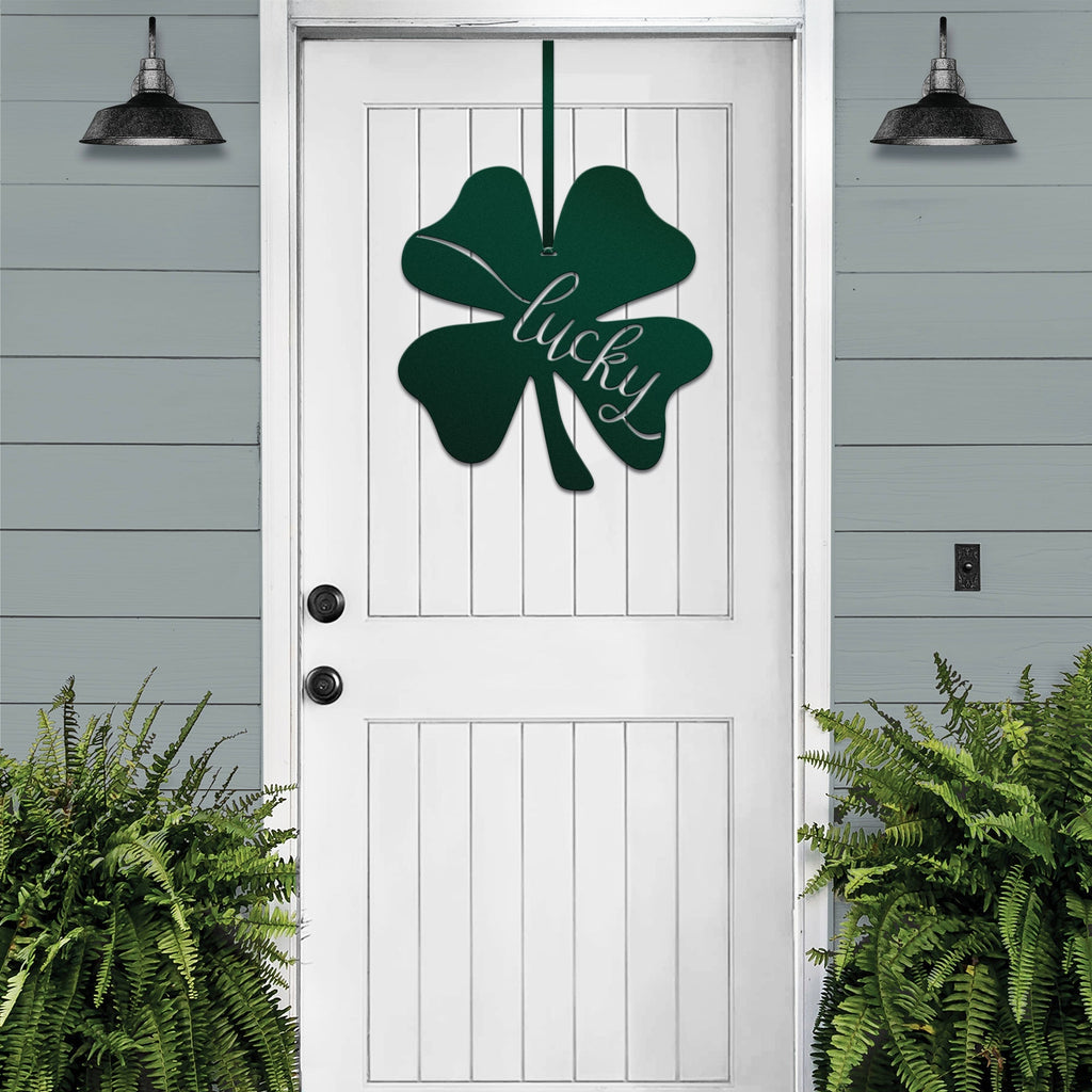 Lucky Shamrocks metal door wreath with green four-leaf clover design and “Lucky” text on white door, festive Irish décor on gray wood wall.