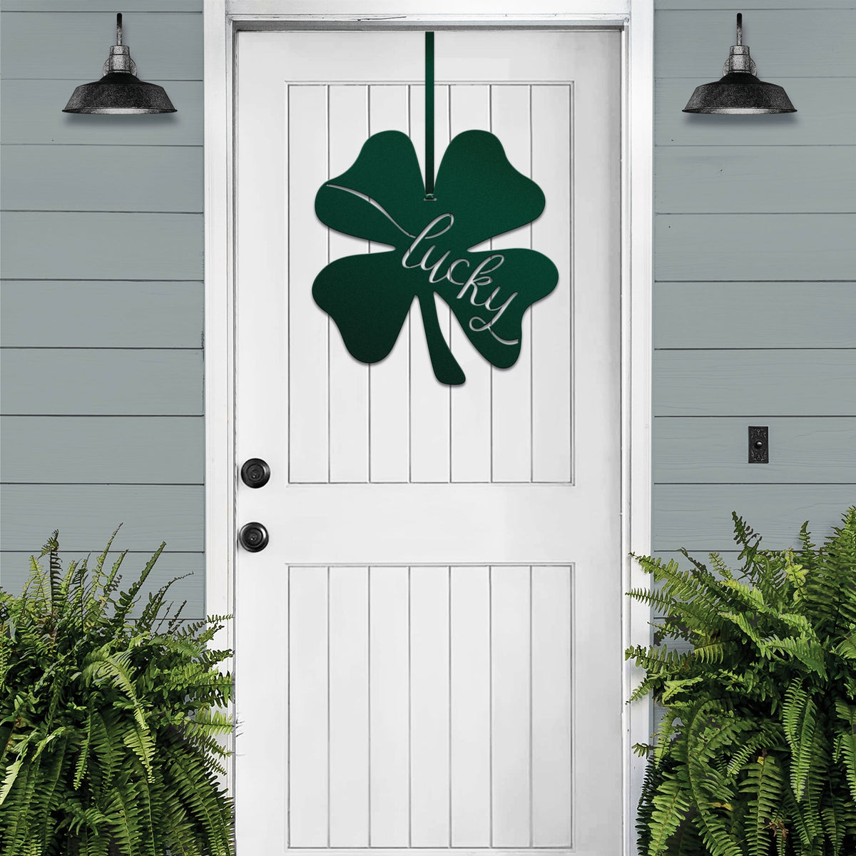 Lucky Shamrocks metal door wreath with green four-leaf clover design and “Lucky” text on white door, festive Irish décor on gray wood wall.