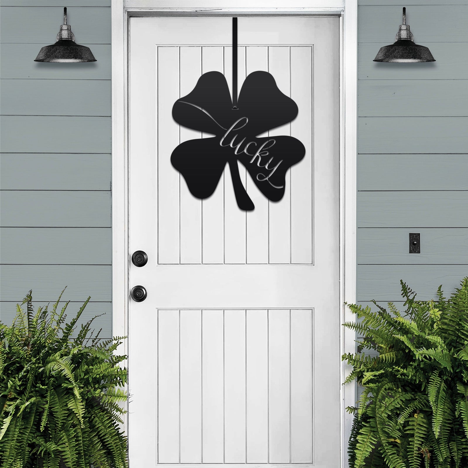 Lucky Shamrocks metal door wreath with black four-leaf clover design and “Lucky” text on white door, festive Irish décor with plants on gray wall.