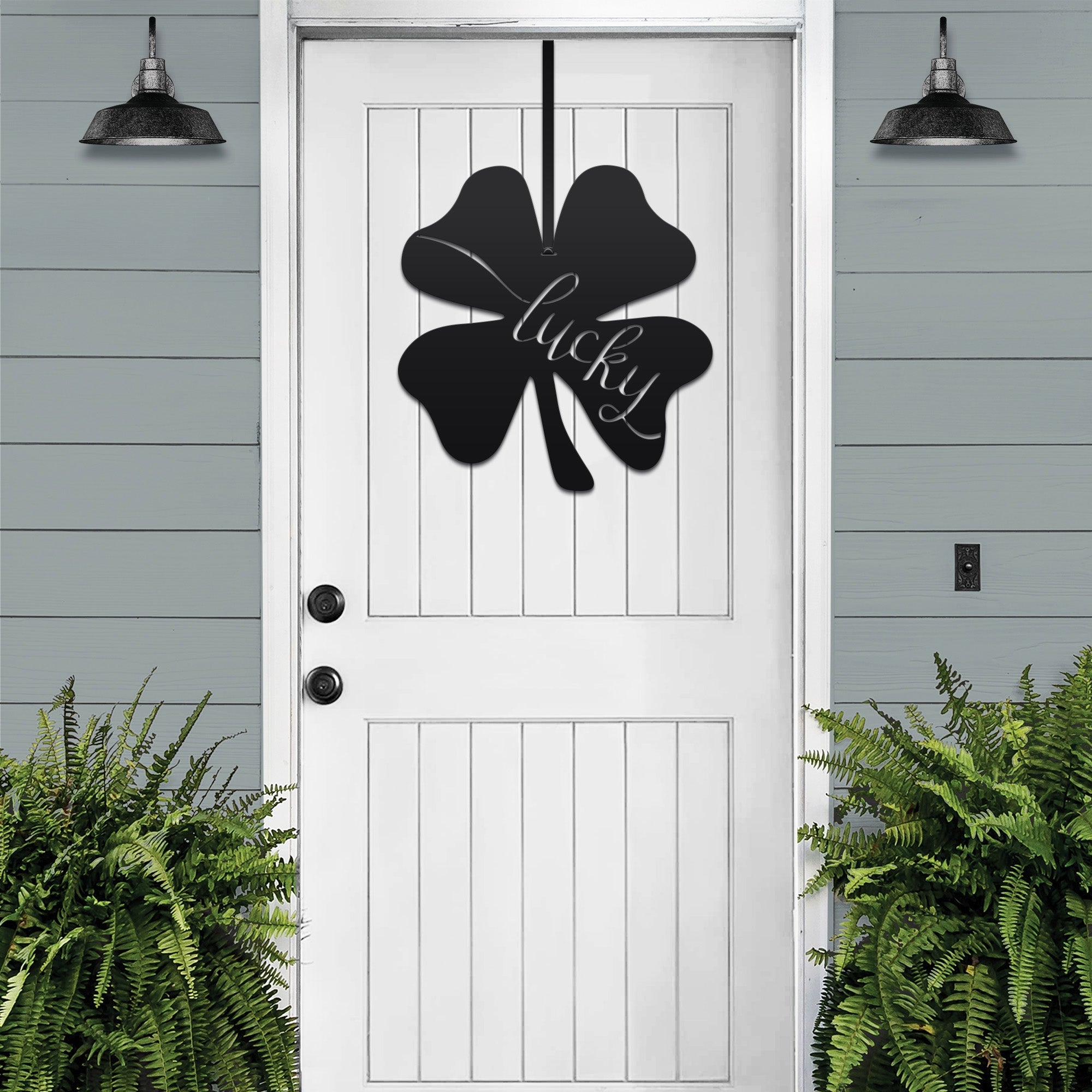Lucky Shamrocks metal door wreath with black four-leaf clover design and “Lucky” text on white door, festive Irish décor with plants on gray wall.