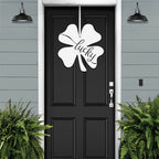 Lucky Shamrocks metal door wreath with white four-leaf clover design and “Lucky” text on black door, festive Irish décor on gray exterior wall.