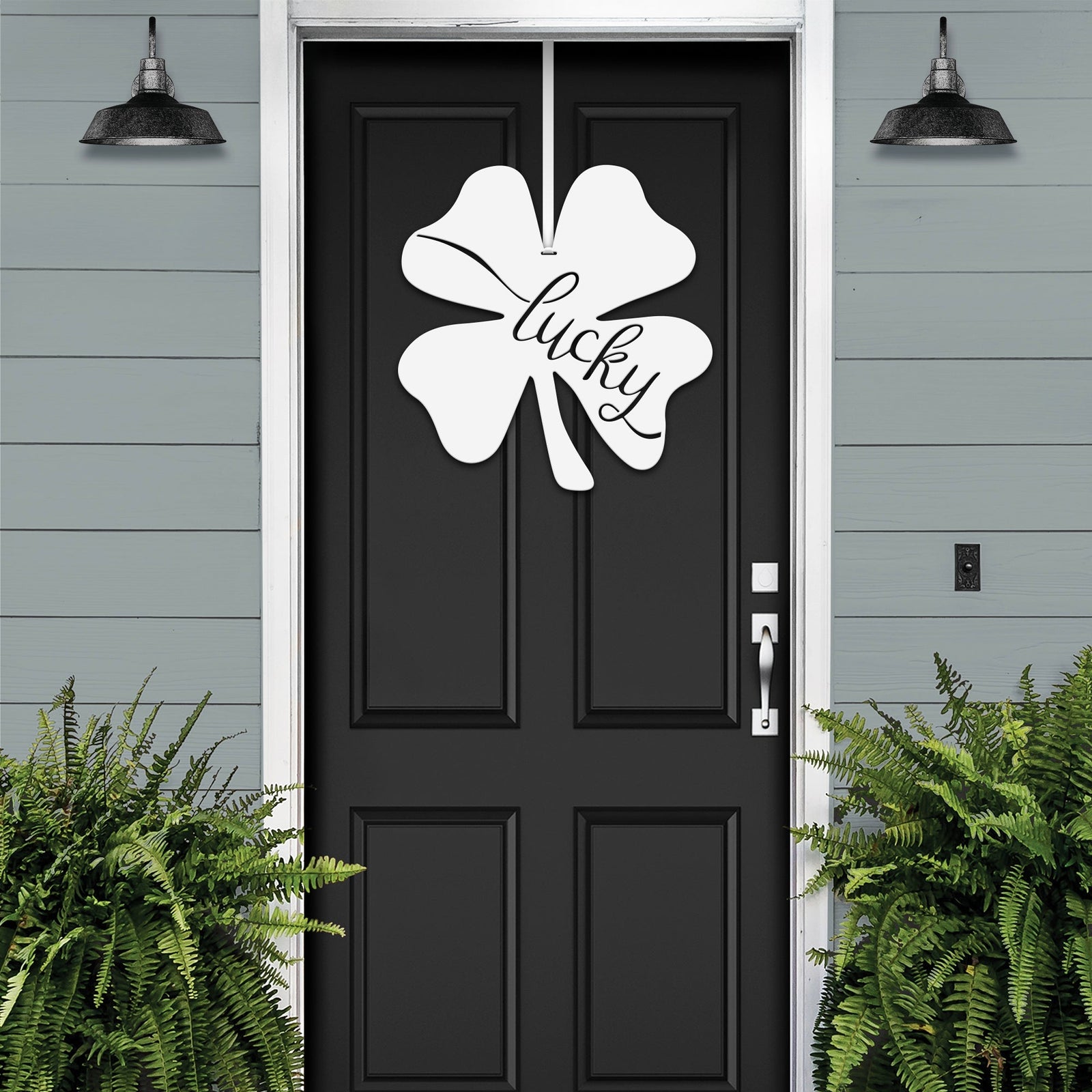 Lucky Shamrocks metal door wreath with white four-leaf clover design and “Lucky” text on black door, festive Irish décor on gray exterior wall.