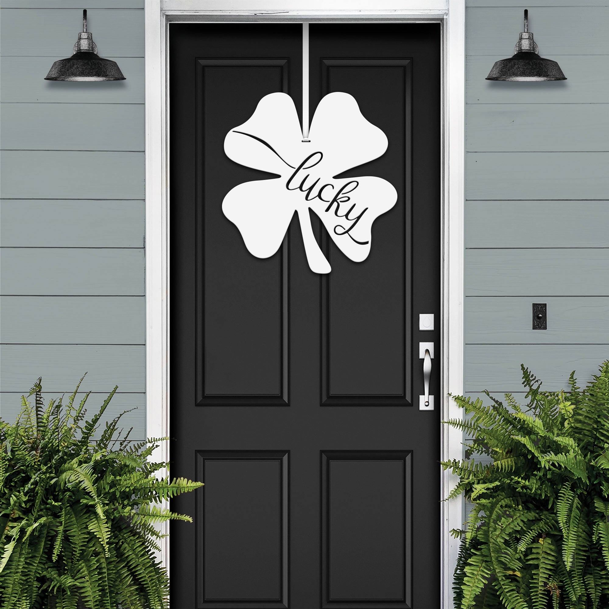 Lucky Shamrocks metal door wreath with white four-leaf clover design and “Lucky” text on black door, festive Irish décor on gray exterior wall.