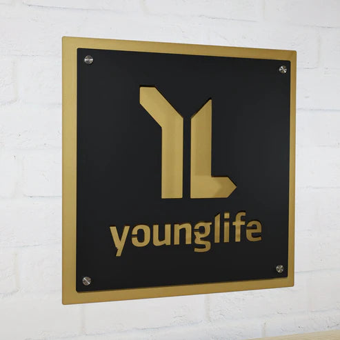 Square metal business sign displaying the "younglife" logo in cutout gold text and design on a matte black faceplate, backed by a gold standoff panel, securely mounted to a white brick wall