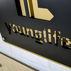 Close-up of a black metal sign featuring a gold “younglife” logo resting on a wooden surface, highlighting elegant design and premium craftsmanship.