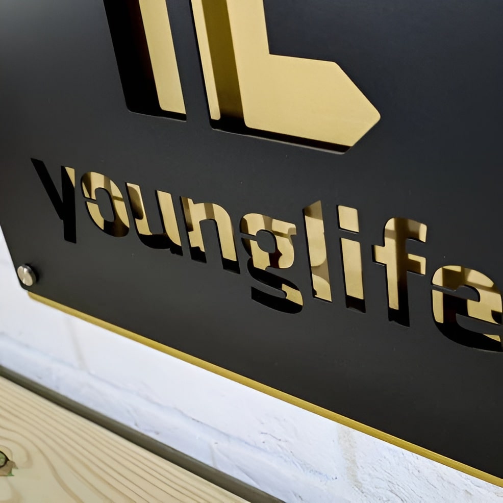 Close-up of a black metal sign featuring a gold “younglife” logo resting on a wooden surface, highlighting elegant design and premium craftsmanship.
