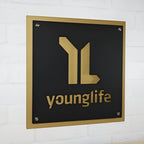 Black metal plaque featuring a gold “younglife” logo mounted on a white brick wall, highlighting a sleek, professional, and elegant design.