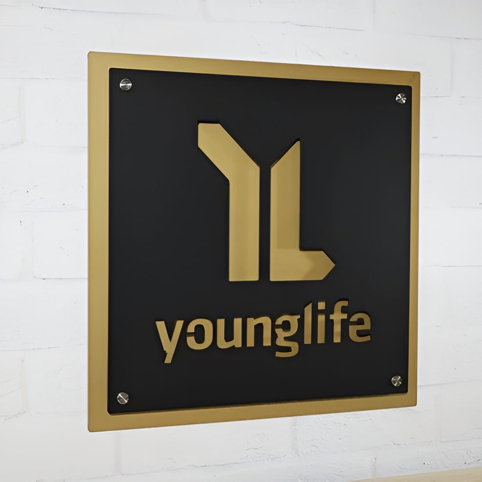 Black metal plaque featuring a gold “younglife” logo mounted on a white brick wall, highlighting a sleek, professional, and elegant design.
