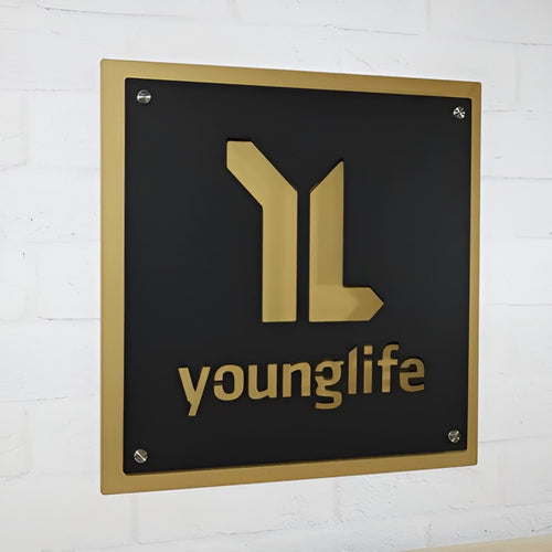 Black metal plaque featuring a gold “younglife” logo mounted on a white brick wall, highlighting a sleek, professional, and elegant design.
