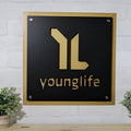 Black metal plaque featuring a gold “younglife” logo mounted on a white brick wall, showcasing a sleek and elegant branded design.