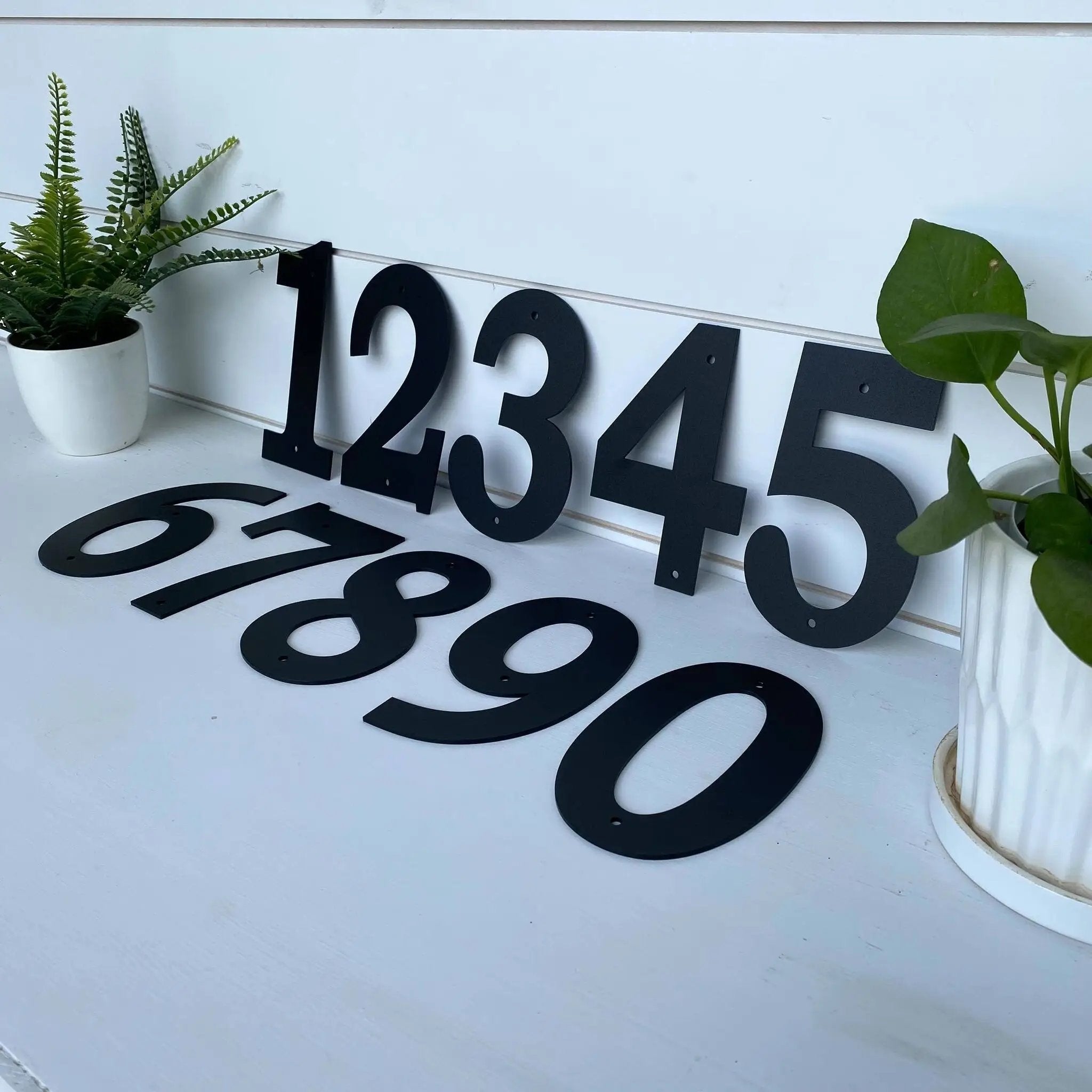 Black house numbers on white wall with plants, modern outdoor address signage, decorative home exterior house number display