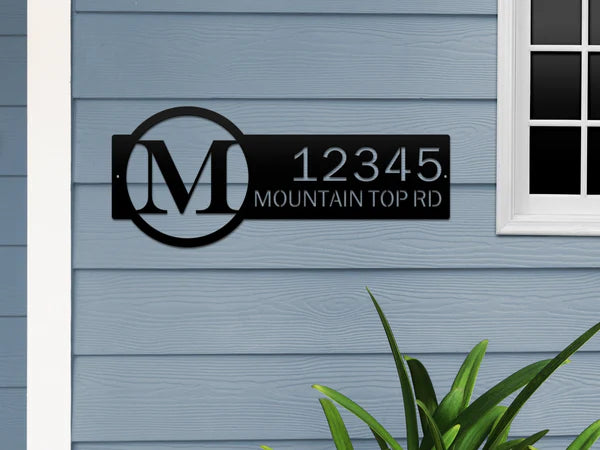 Black metal monogram sign with letter 'M' and '12345 Mountain Top RD', on bluish-gray wall beside a door.