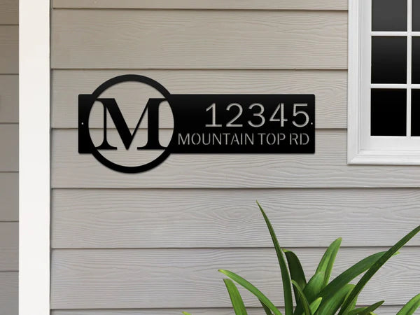 Black metal monogram sign with letter 'M' and '12345 Mountain Top RD', on beige wall beside a door.