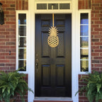HouseSensationsArt metal over-the-door wreath hook in gold pineapple design, displayed on black front door of brick house, part of 90-color USA-made collection.