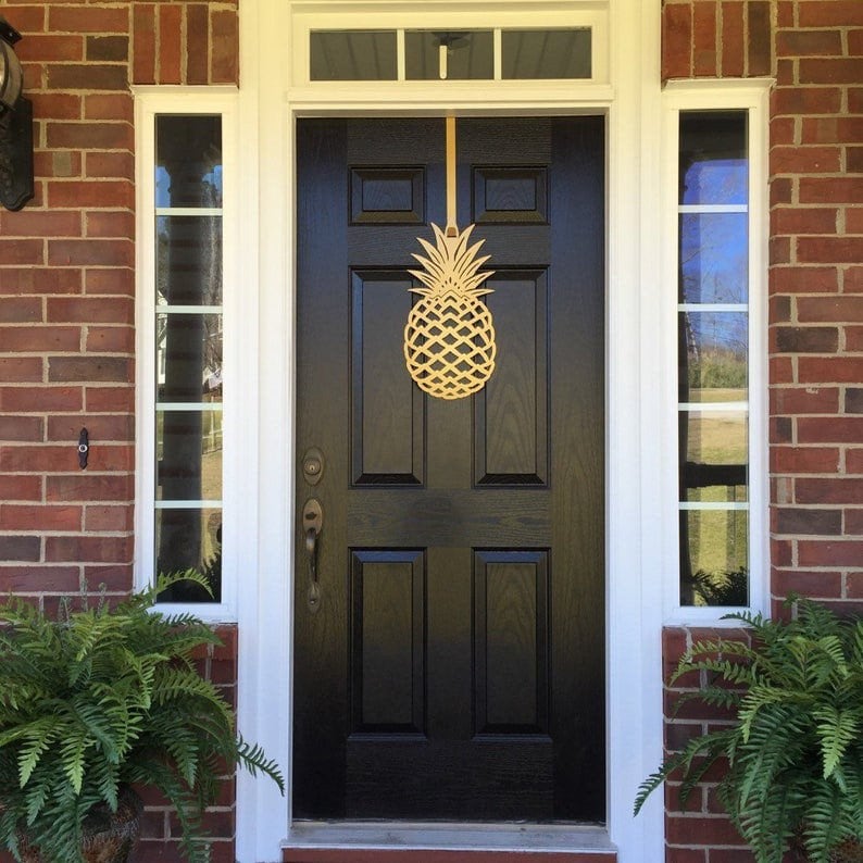 HouseSensationsArt metal over-the-door wreath hook in gold pineapple design, displayed on black front door of brick house, part of 90-color USA-made collection.