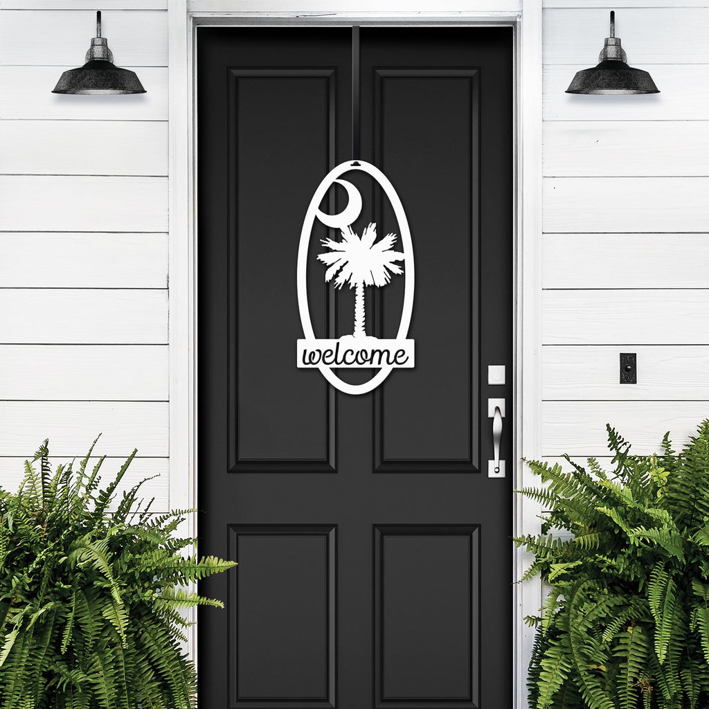 Black front door with decorative white palm tree “Welcome” decal, flanked by green plants on white wall, custom outdoor home entrance décor