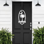 Black front door with decorative white palm tree “Welcome” decal, flanked by green plants on white wall, custom outdoor home entrance décor