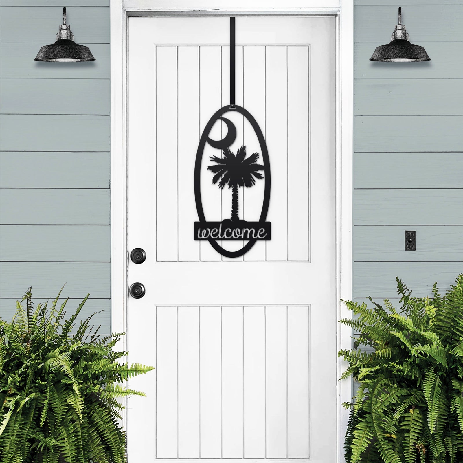 White door with decorative black “Welcome” sign featuring palm tree design, flanked by green plants against light blue wall, custom outdoor home entrance décor