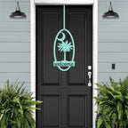 Black door with decorative palm tree “Welcome” decal, flanked by green plants against light blue wall, custom outdoor home entrance décor
