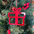 Red gift-shaped Christmas ornament hanging on a tree with green leaves and red berries, festive holiday decoration.