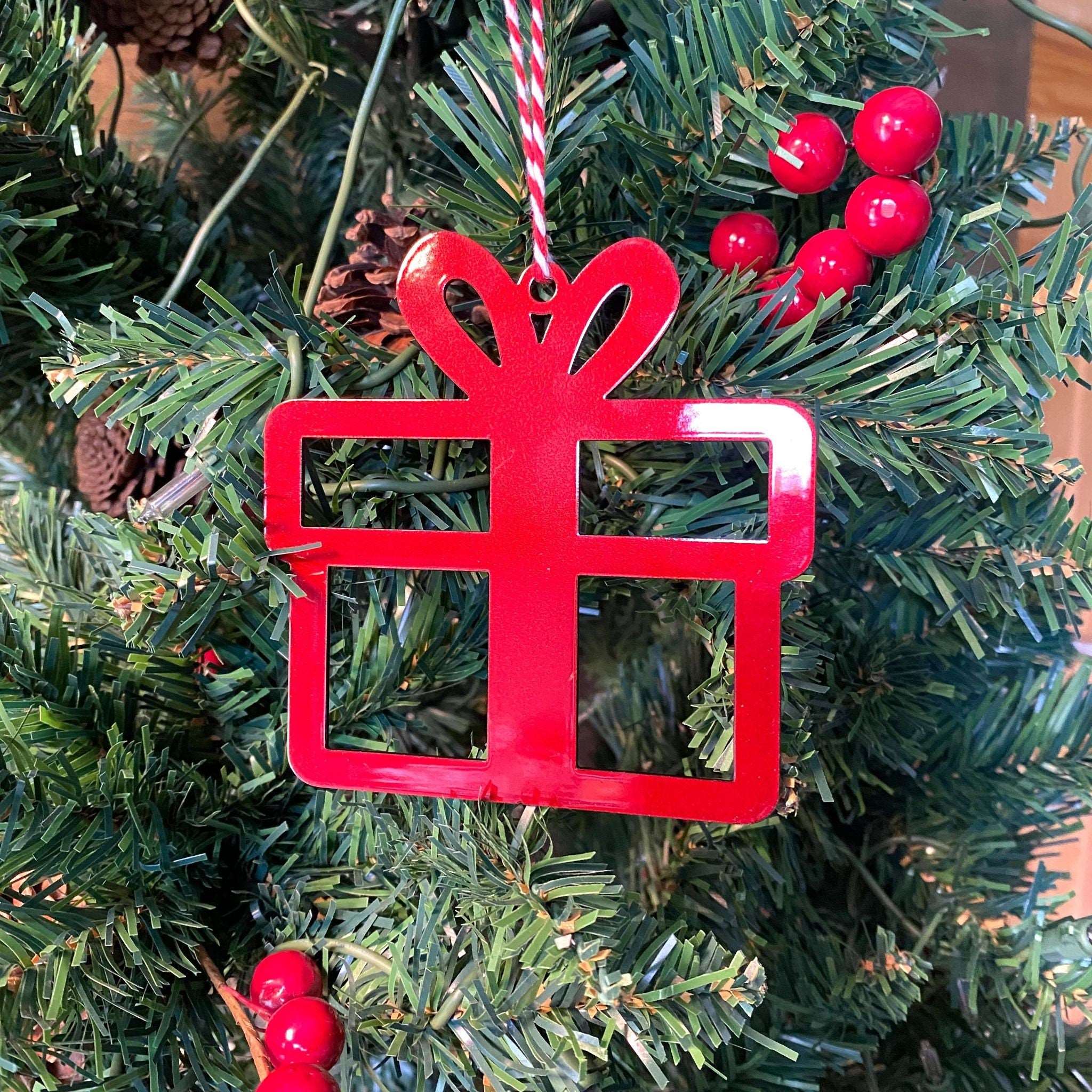 Red gift-shaped Christmas ornament hanging on a tree with green leaves and red berries, festive holiday decoration.