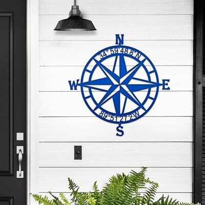Custom nautical compass rose metal wall art in blue mounted on white wooden wall with black door and green plants, coastal décor, HouseSensationsArt