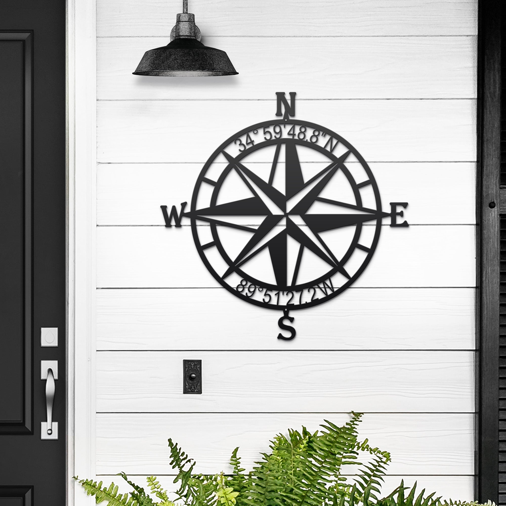 Black nautical compass rose metal wall décor mounted on white wooden wall with green plants at the bottom, coastal-inspired décor, HouseSensationsArt