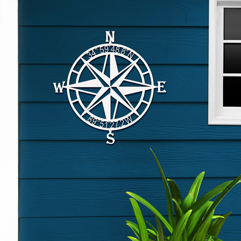 White nautical compass rose metal wall art mounted on blue wooden wall with a plant in foreground, coastal décor, HouseSensationsArt