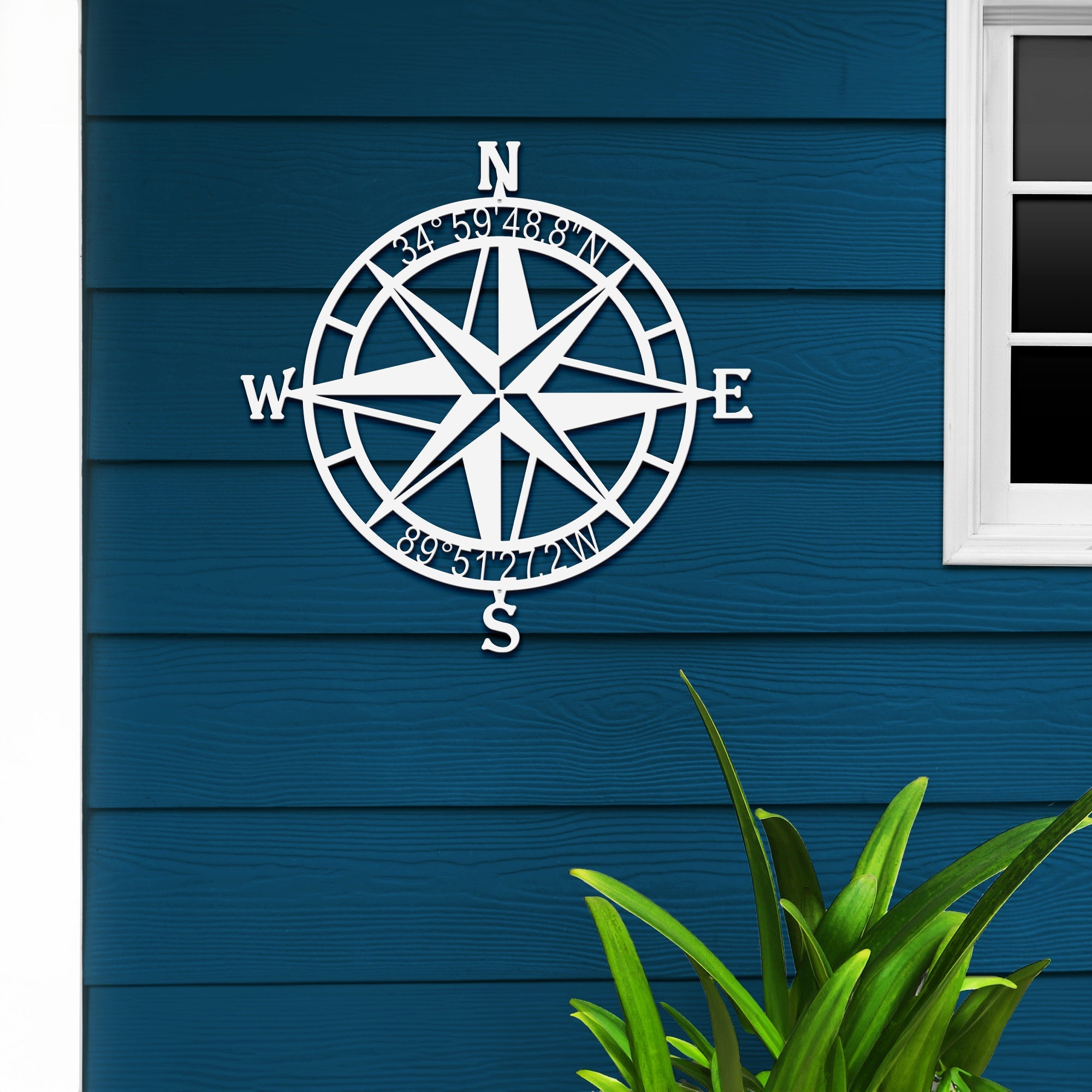 White nautical compass rose metal wall art mounted on blue wooden wall with a plant in foreground, coastal décor, HouseSensationsArt