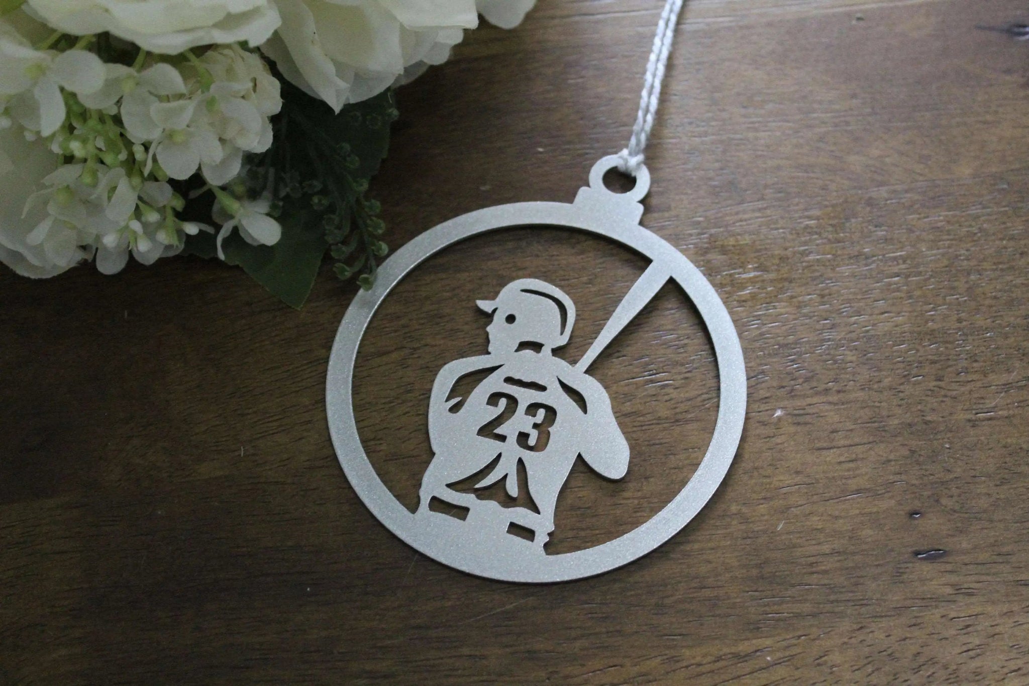 Metallic pendant featuring design of person holding number 23, displayed on wooden surface with flowers, decorative keepsake or commemorative ornament for gifts and seasonal décor