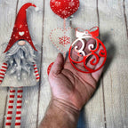 Hand holding red decorative ornament with Christmas-themed background featuring gnome and snowflakes, festive holiday decoration for seasonal décor and gift display