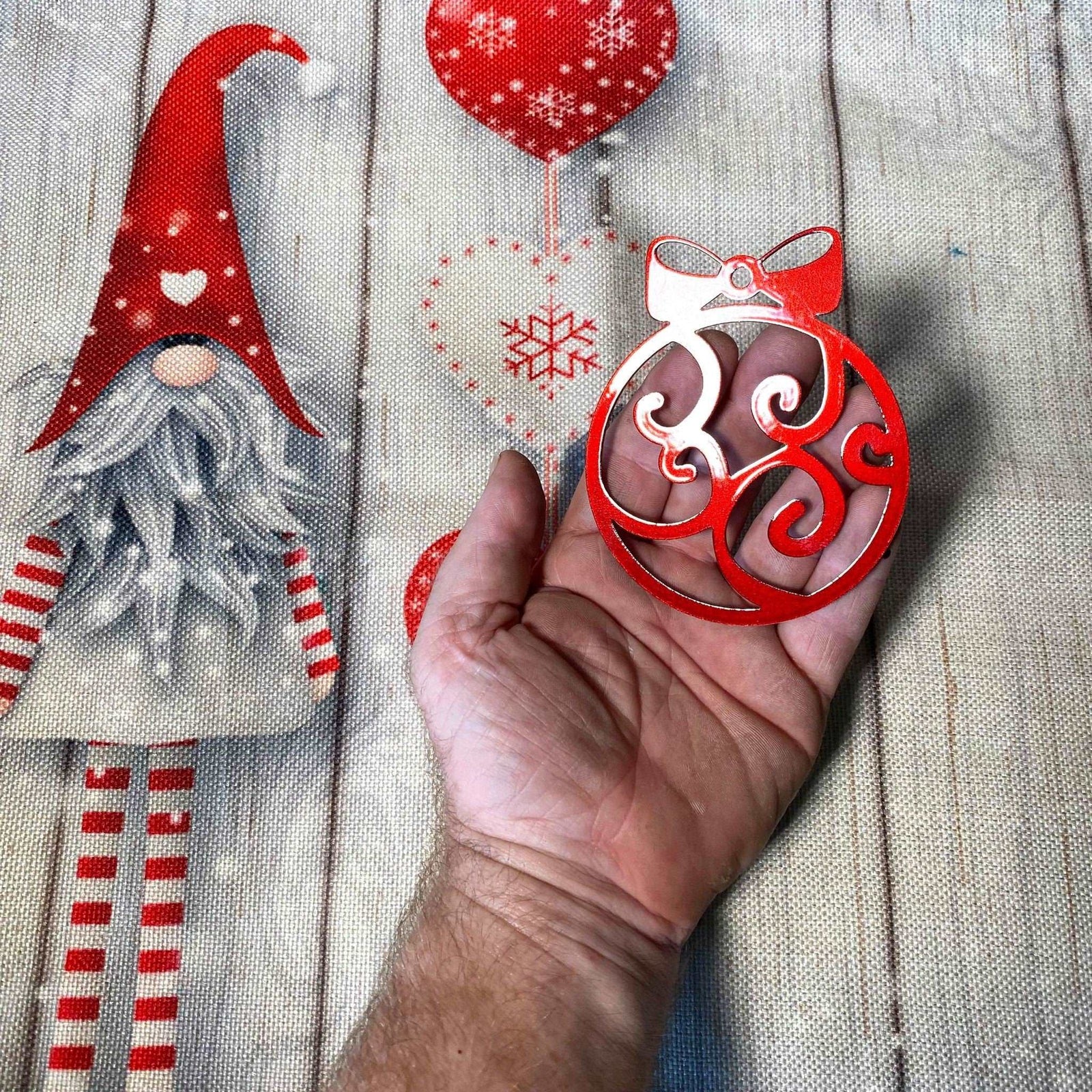 Hand holding red decorative ornament with Christmas-themed background featuring gnome and snowflakes, festive holiday decoration for seasonal décor and gift display