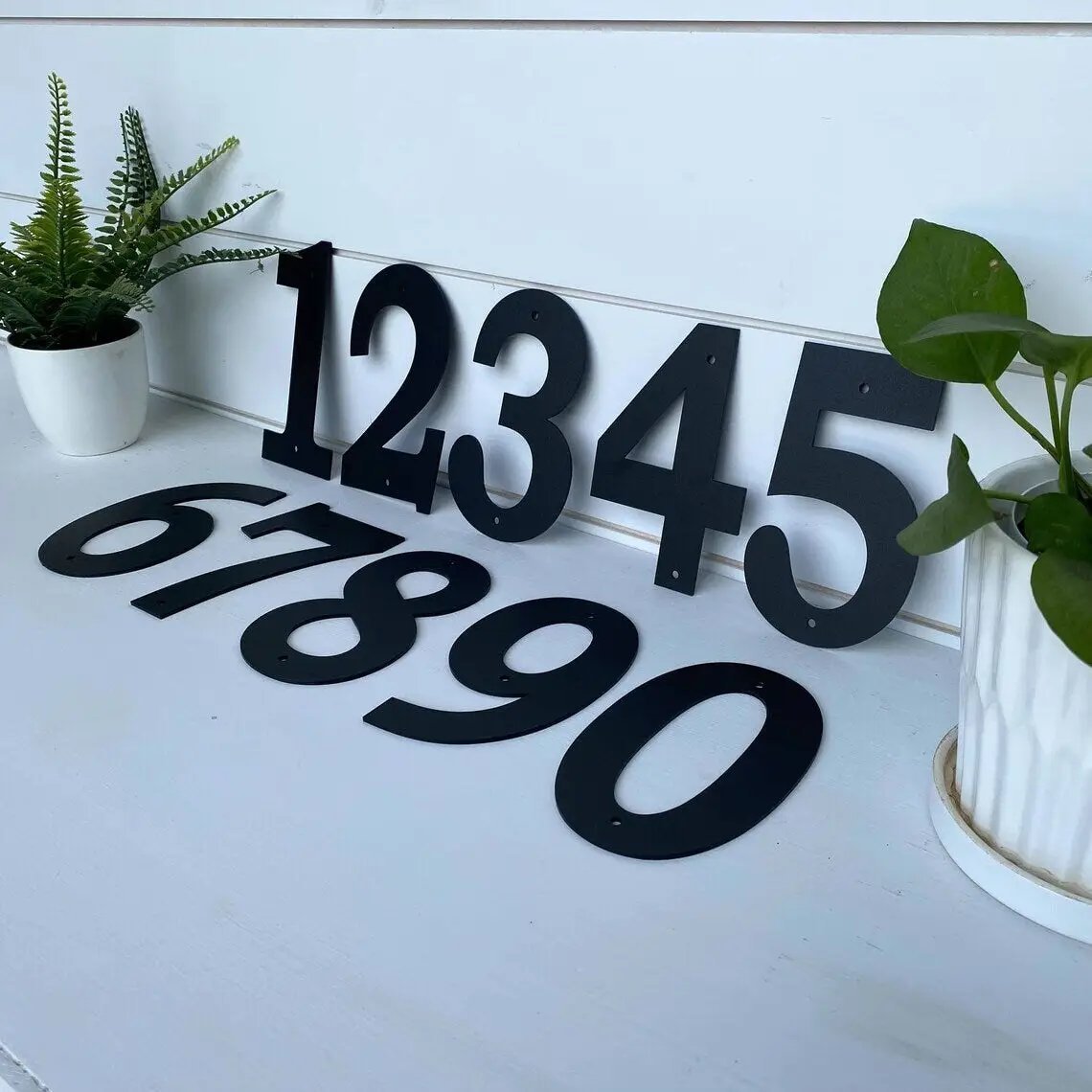 Black house numbers on white wooden surface with plants for custom outdoor address signs, personalized front door numbers, and decorative curb appeal