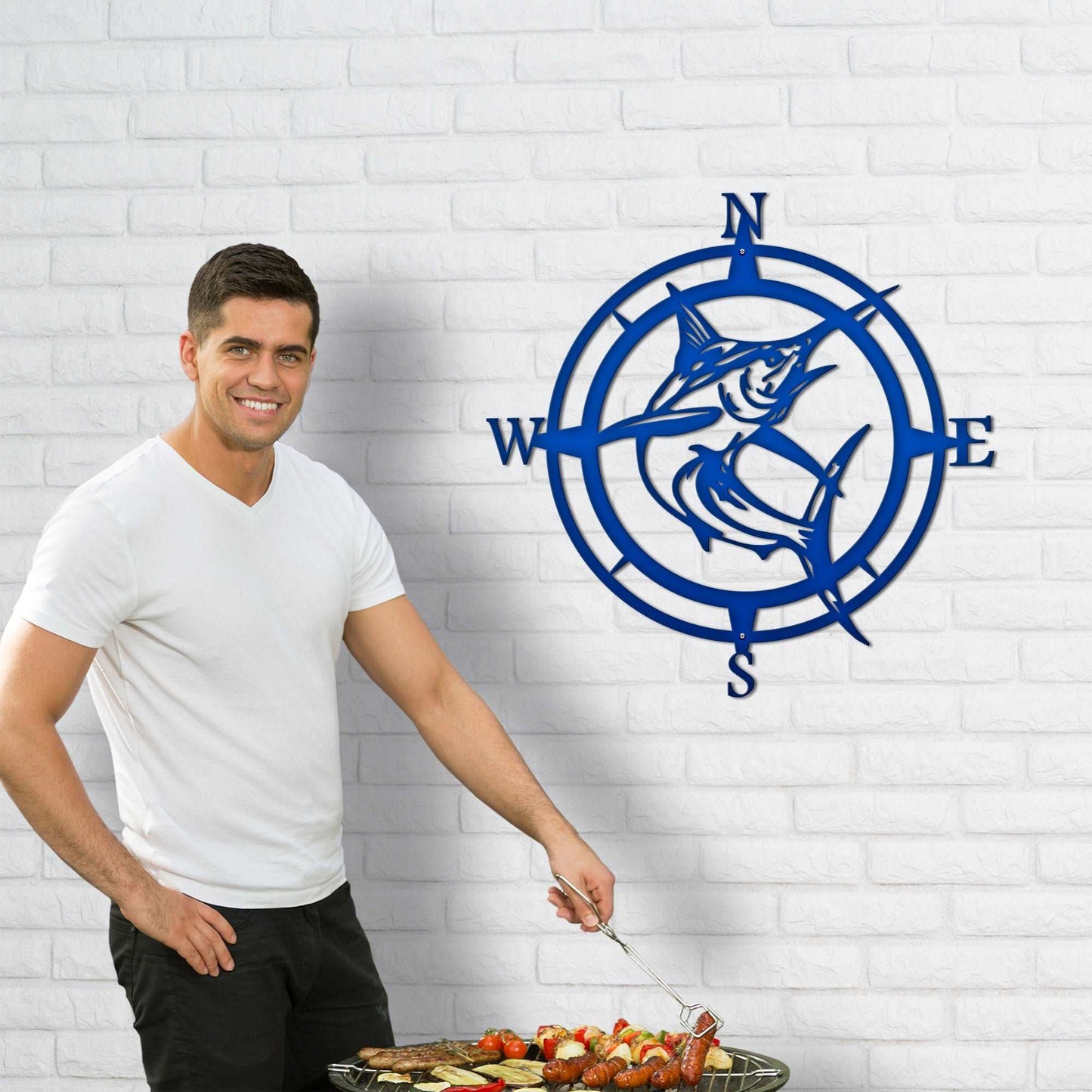 Man grilling outdoors next to decorative metal wall art with fish design on white brick wall, nautical-themed outdoor décor, custom home exterior accent