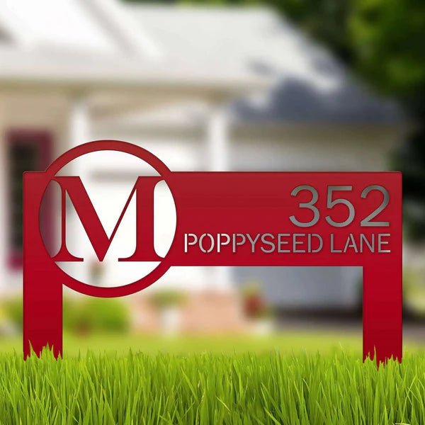 Red metal sign displaying an “M” logo and house number “352” positioned on grass in front of a blurred house, showcasing durable outdoor design and clear numbering.